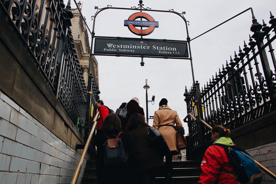 A group of people ascending outdoor stairs at Westminster Underground Station, with a black wrought iron fence on either side and a sign overhead indicating the station's name and its connection to the London Underground. The group includes individuals wearing jackets in various colours, some carrying backpacks and shoulder bags. The stairs are made of dark material, possibly stone or concrete, leading up to the station entrance, which is situated beneath a large, curved sign with the underground logo. The environment suggests an urban setting with overcast sky and street lamps visible in the background, consistent with a typical busy London station entrance, relevant to home relocation or moving services such as those offered by Streatham Movers.