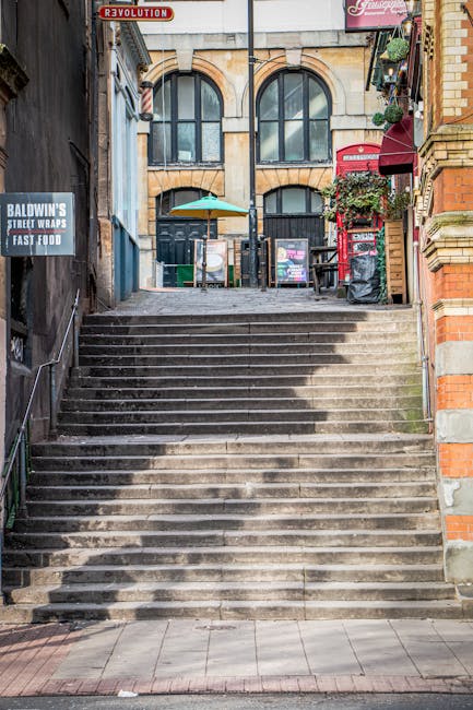 A wide exterior view of a set of concrete stairs leading up to a building entrance on a narrow street, with black metal handrails along both sides. The stairs are weathered and uneven, with some visible stains and dirt. At the top, there is a stone facade with large arched windows and a dark doorway in the center. To the left, a sign advertises Baldwin's Street Wraps and Fast Food, mounted on a dark brick wall, while to the right, a red British telephone booth and a small potted plant with flowers are attached to a brick building. The scene is illuminated by natural daylight, capturing the urban environment typical of London, suitable for a house or flat relocation context involving stair navigation. Streatham Movers specialises in home removals and furniture transport, often dealing with tight staircases and limited access, as demonstrated by this setting.