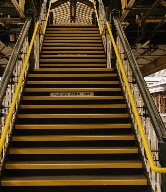 A group of people ascending outdoor stairs at Westminster Underground Station, with a black wrought iron fence on either side and a sign overhead indicating the station's name and its connection to the London Underground. The group includes individuals wearing jackets in various colours, some carrying backpacks and shoulder bags. The stairs are made of dark material, possibly stone or concrete, leading up to the station entrance, which is situated beneath a large, curved sign with the underground logo. The environment suggests an urban setting with overcast sky and street lamps visible in the background, consistent with a typical busy London station entrance, relevant to home relocation or moving services such as those offered by Streatham Movers.
